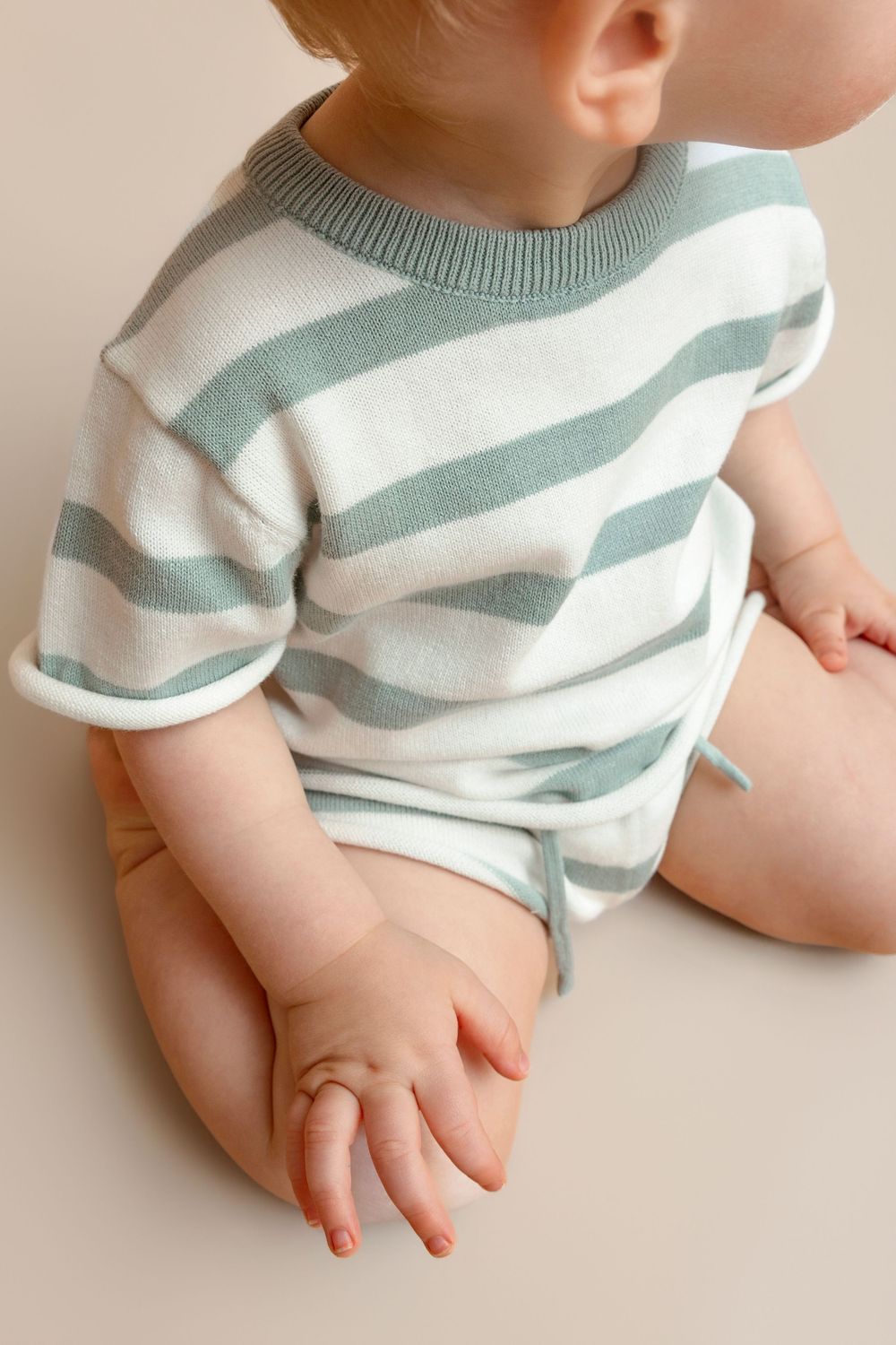 Baby wearing a sage green and white striped knit onesie, sitting on a beige background.