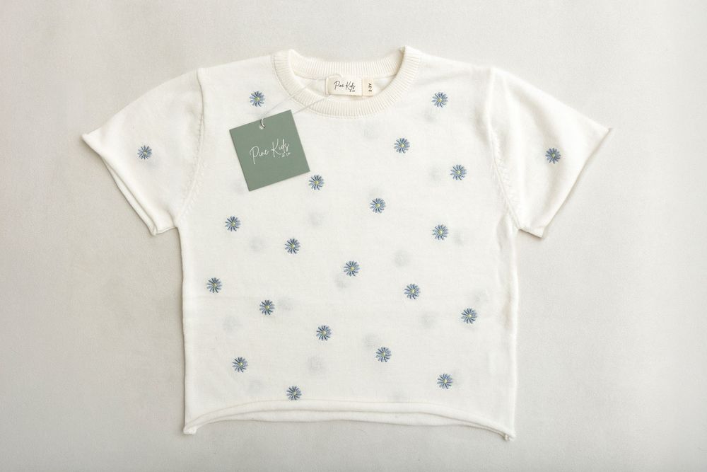 White short-sleeve kids knit top with blue polka dot pattern and green brand tag, laid flat on gray background.