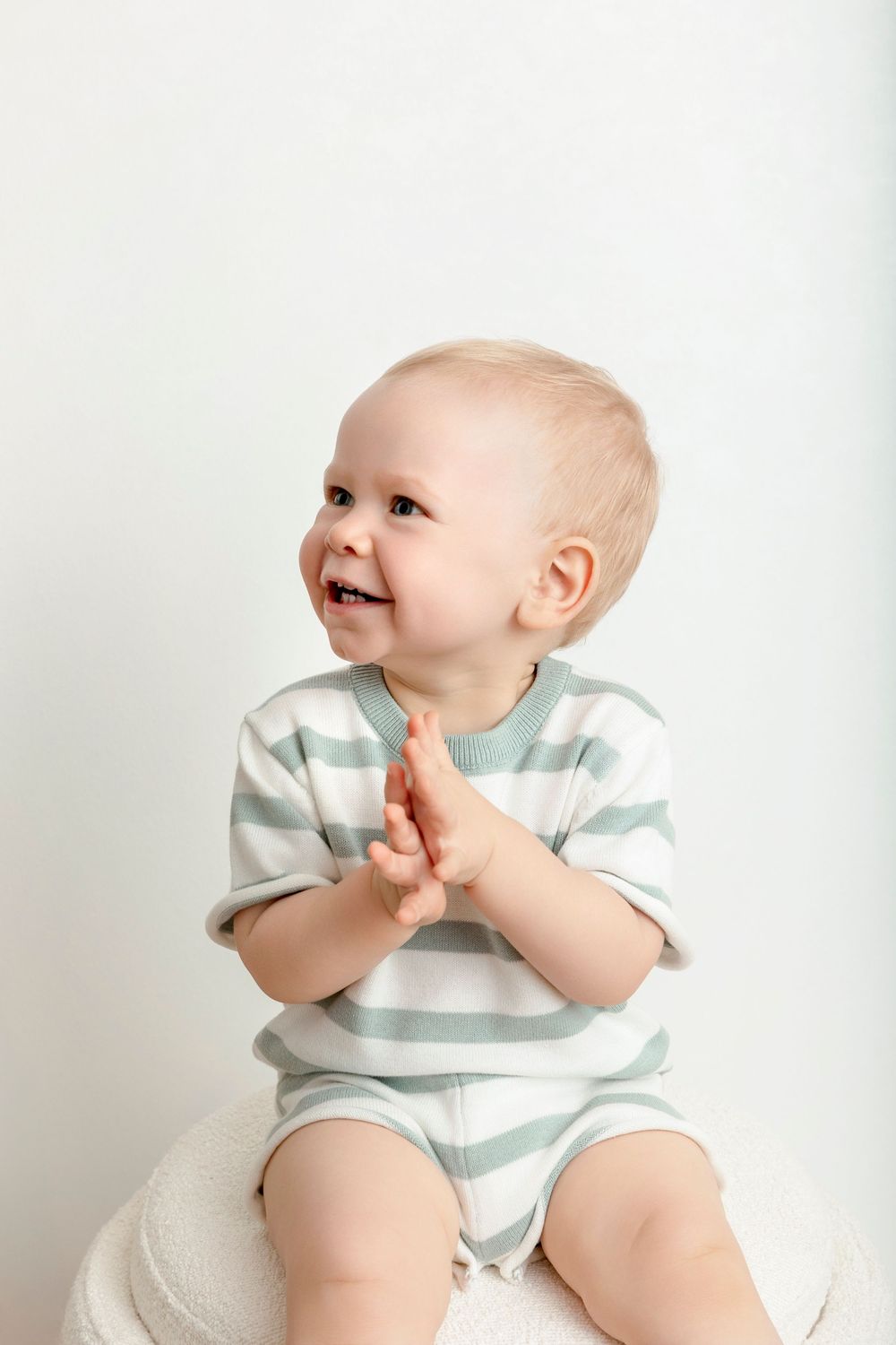 Smiling baby in striped romper clapping hands, sitting on white surface against light background.