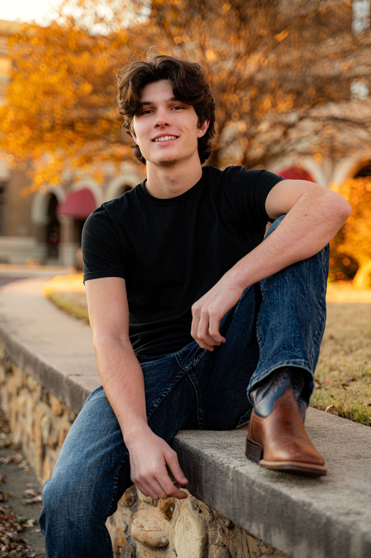 Young man in black t-shirt and jeans sitting on stone wall outdoors with autumn foliage background.