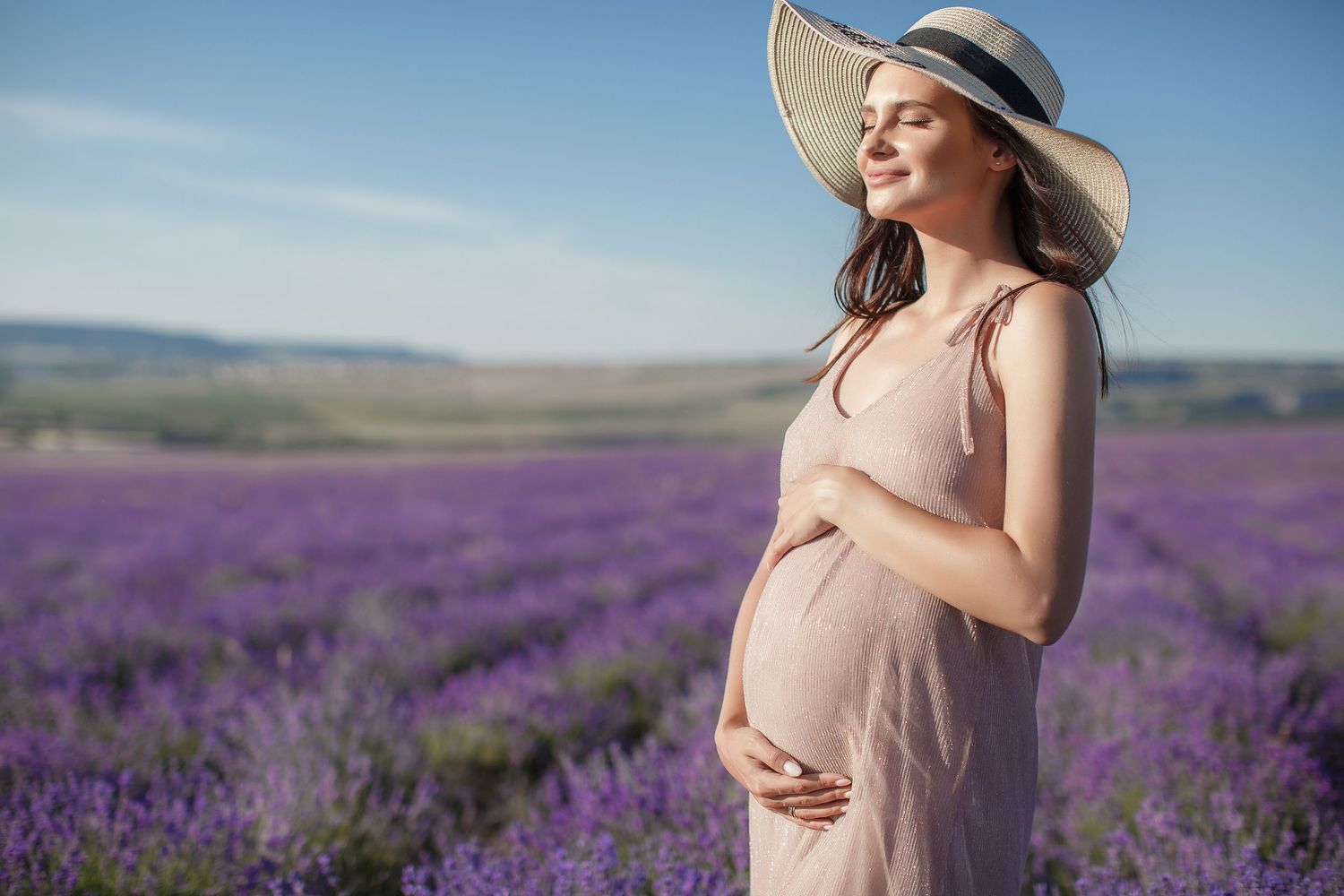 Pregnant woman in pink dress and sun hat cradling her belly in a vibrant purple lavender field on a sunny day.