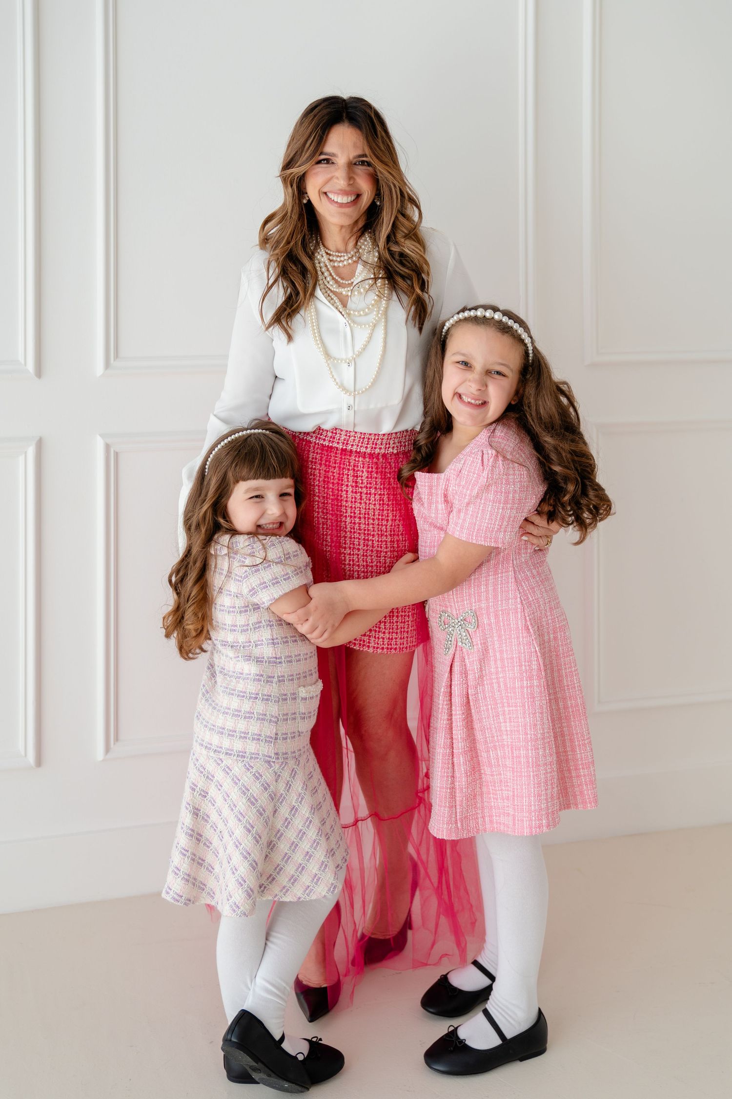 A woman in pink tweed skirt and white blouse poses with two young girls in matching pink dresses in a bright studio.
