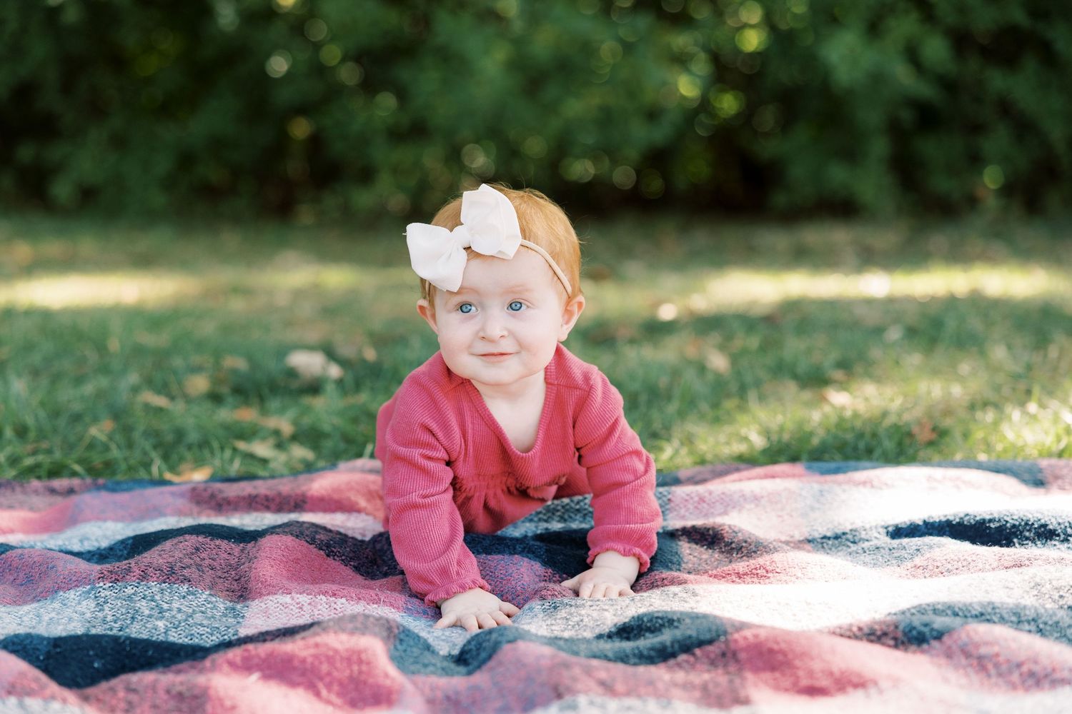 Baby girl in pink outfit with white bow headband crawling on plaid blanket in park.