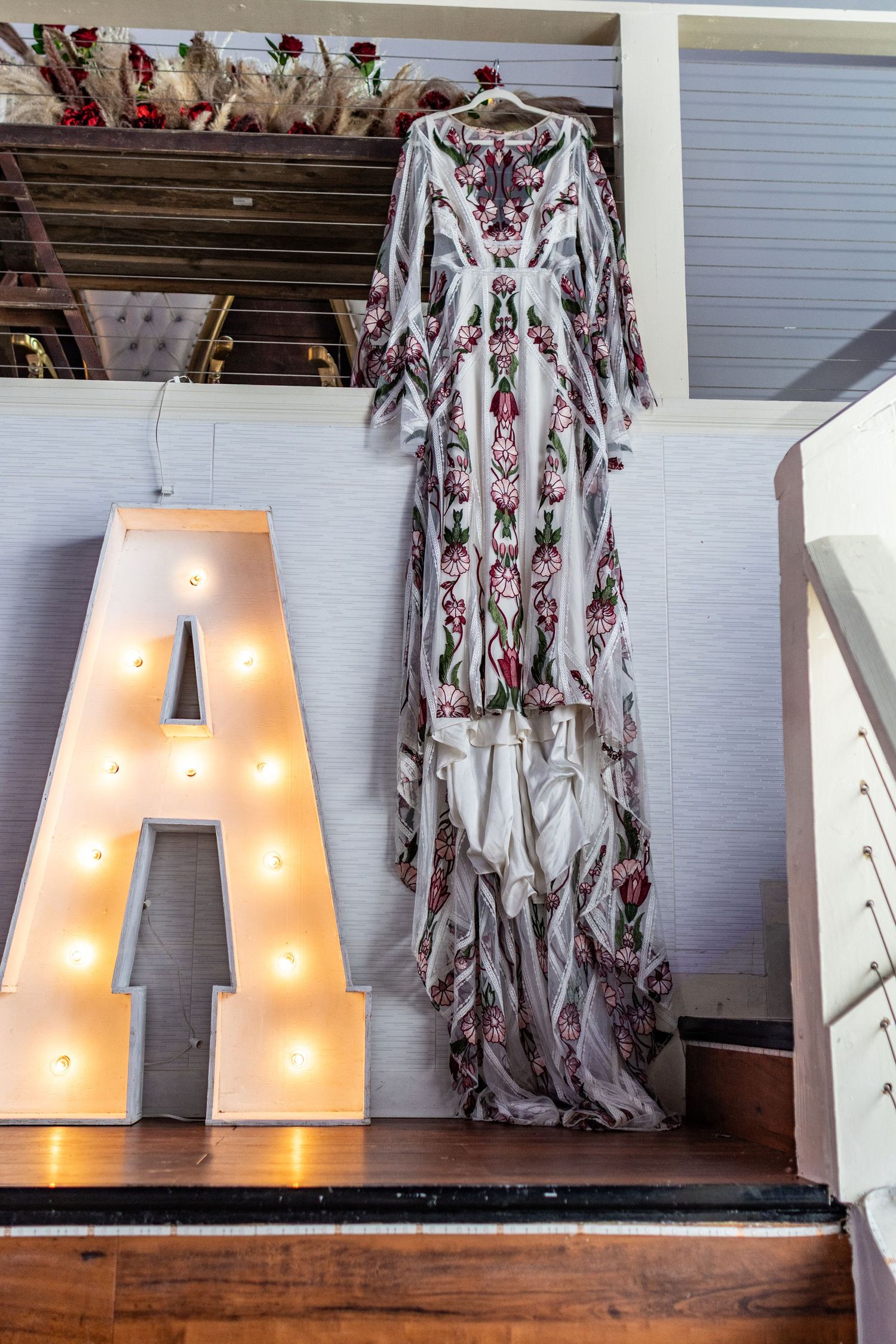 Illuminated marquee letter A beside a floral boho maxi dress hanging on display in a rustic venue.