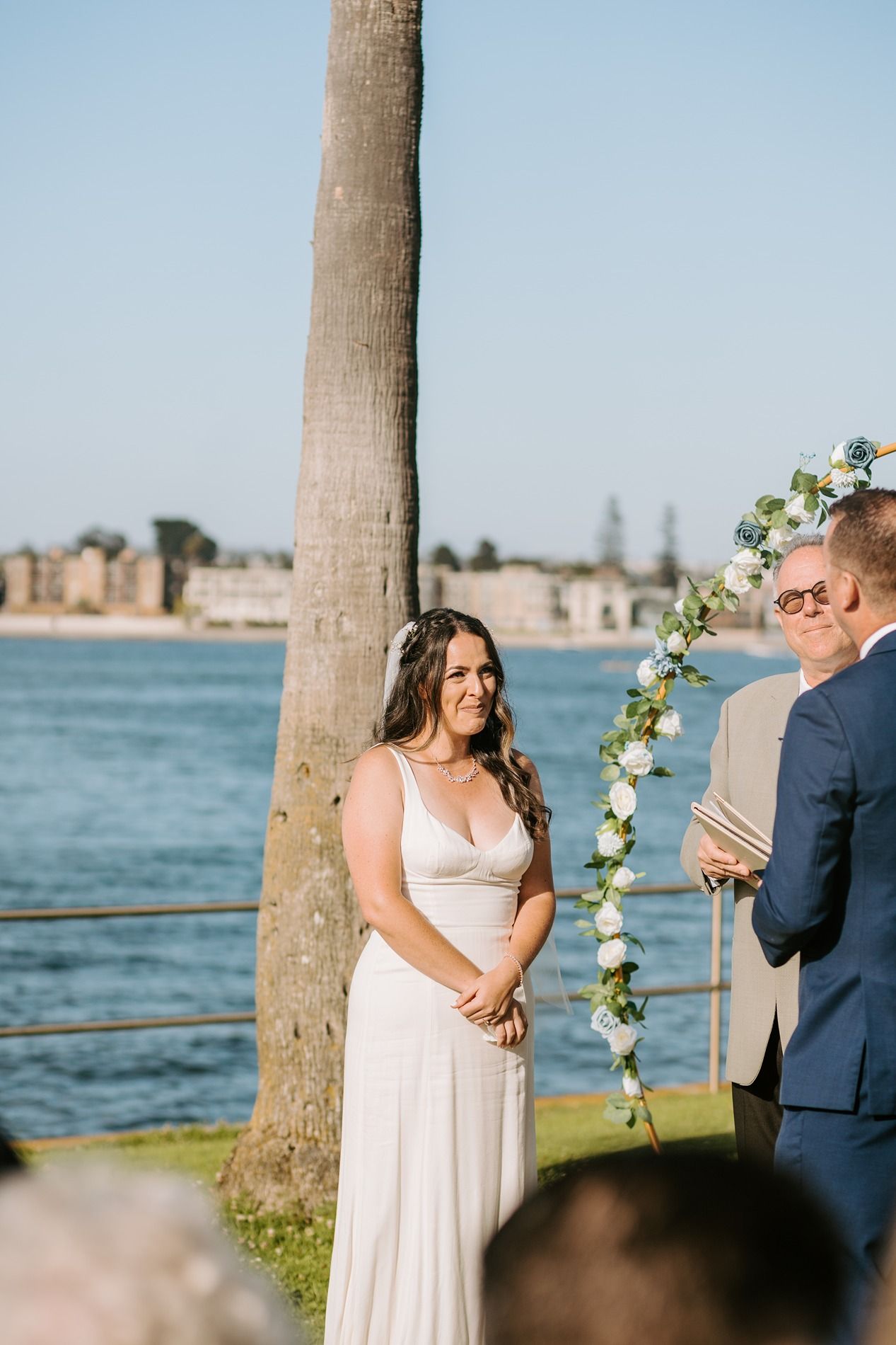 Bride smiling at groom during outdoor waterfront wedding ceremony with floral arch and palm tree at Tower Beach Club.