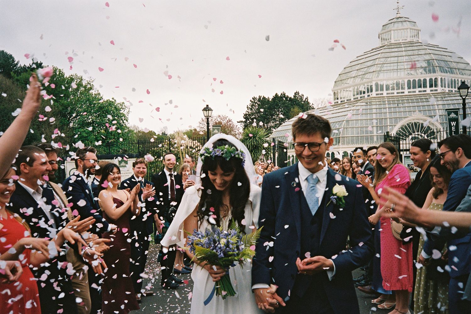 Newlywed couple walks through confetti shower outside a glasshouse surrounded by cheering wedding guests. Shot on 35mm film.