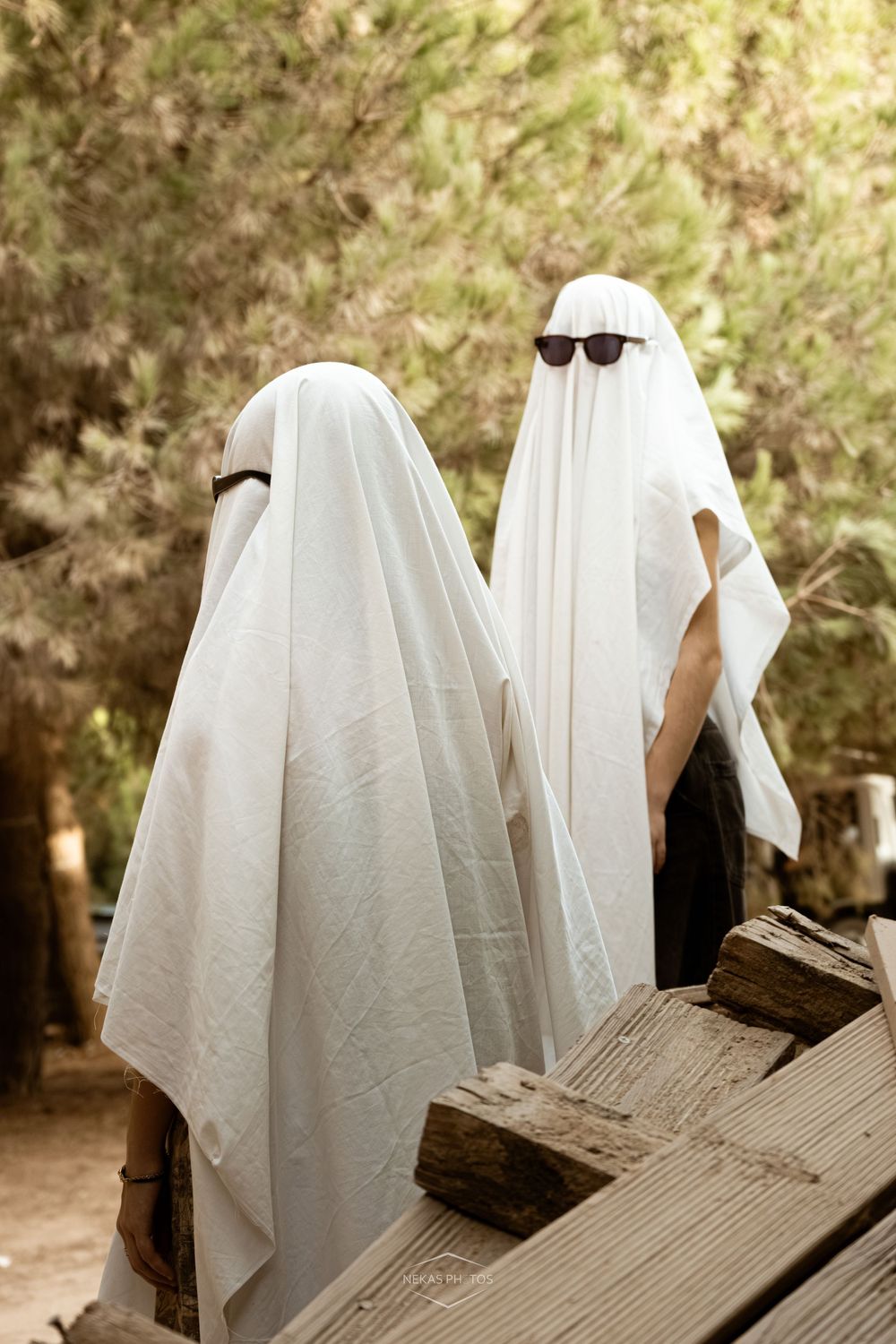 Two people dressed as ghosts in white sheets with sunglasses stand near wooden fence outdoors.