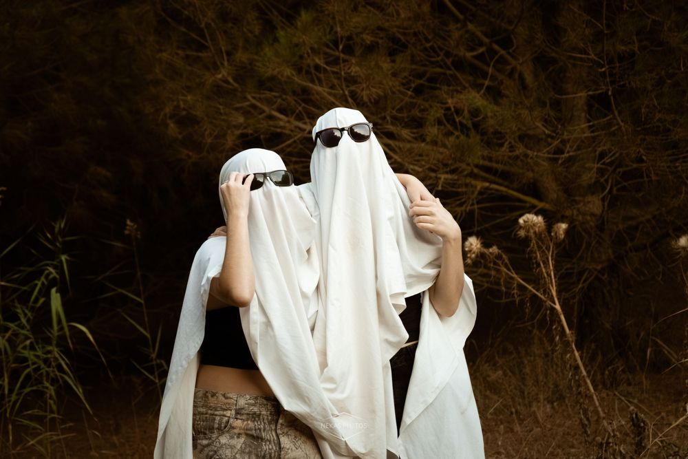 Two people dressed as ghosts in white sheets with sunglasses pose outdoors near trees.