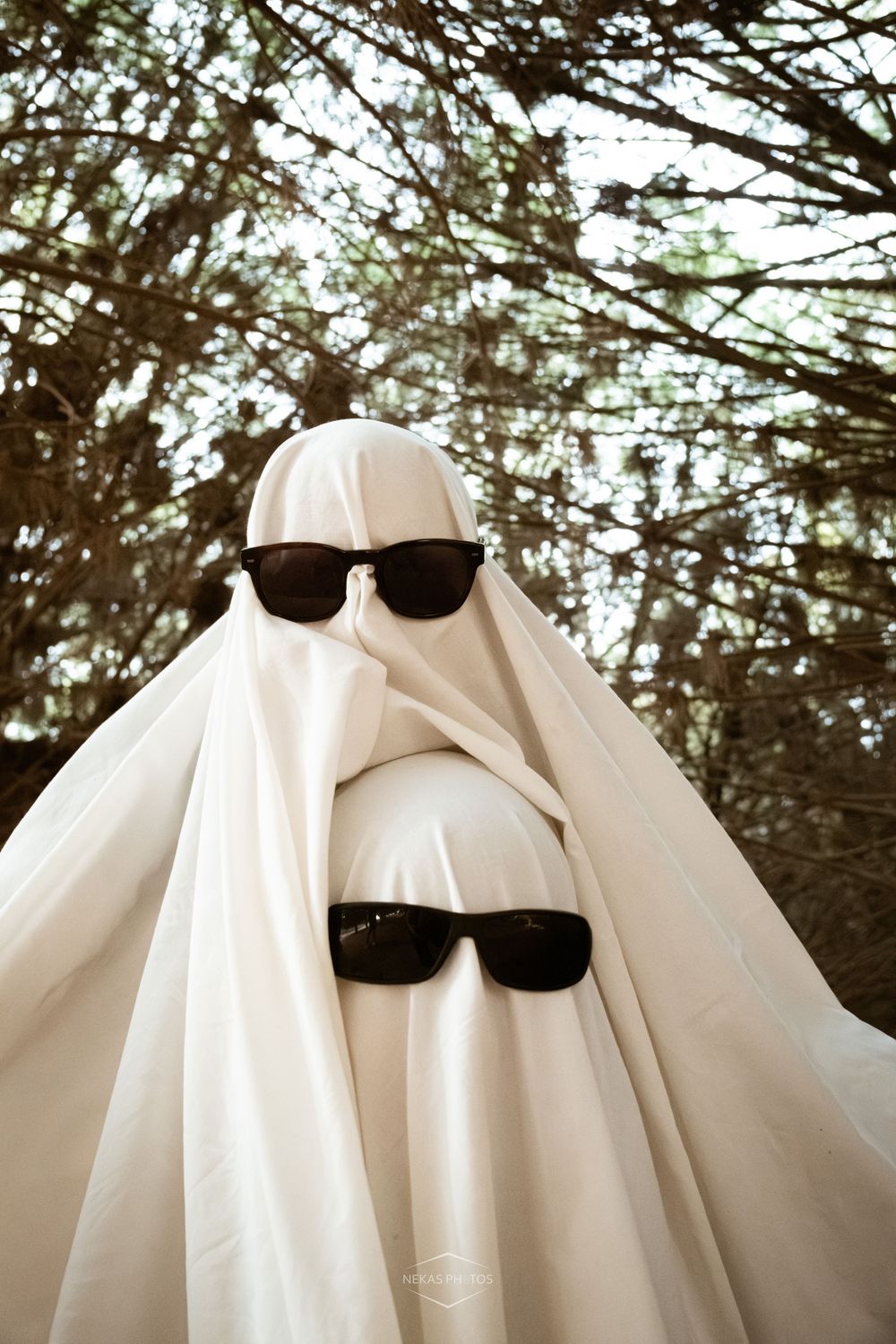 Two sunglasses worn by a person dressed as a ghost in a white sheet costume standing among trees.