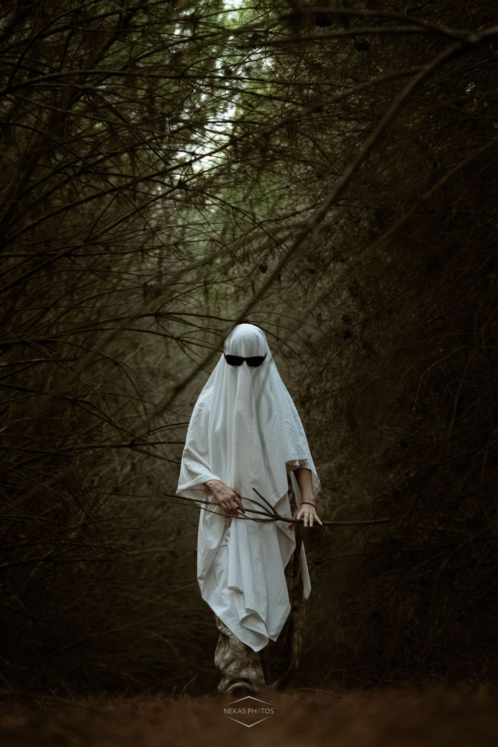 Person dressed as a ghost in white sheet with sunglasses holding a stick in a dark forest path.