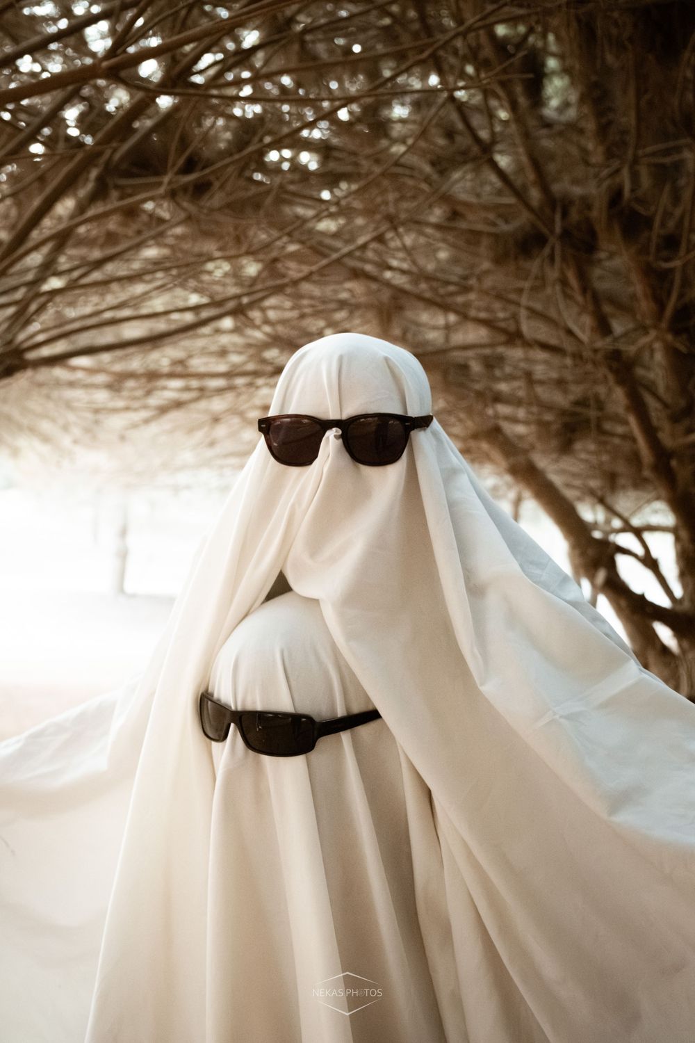 Person dressed as a ghost in white sheet with sunglasses poses outdoors under bare winter trees.