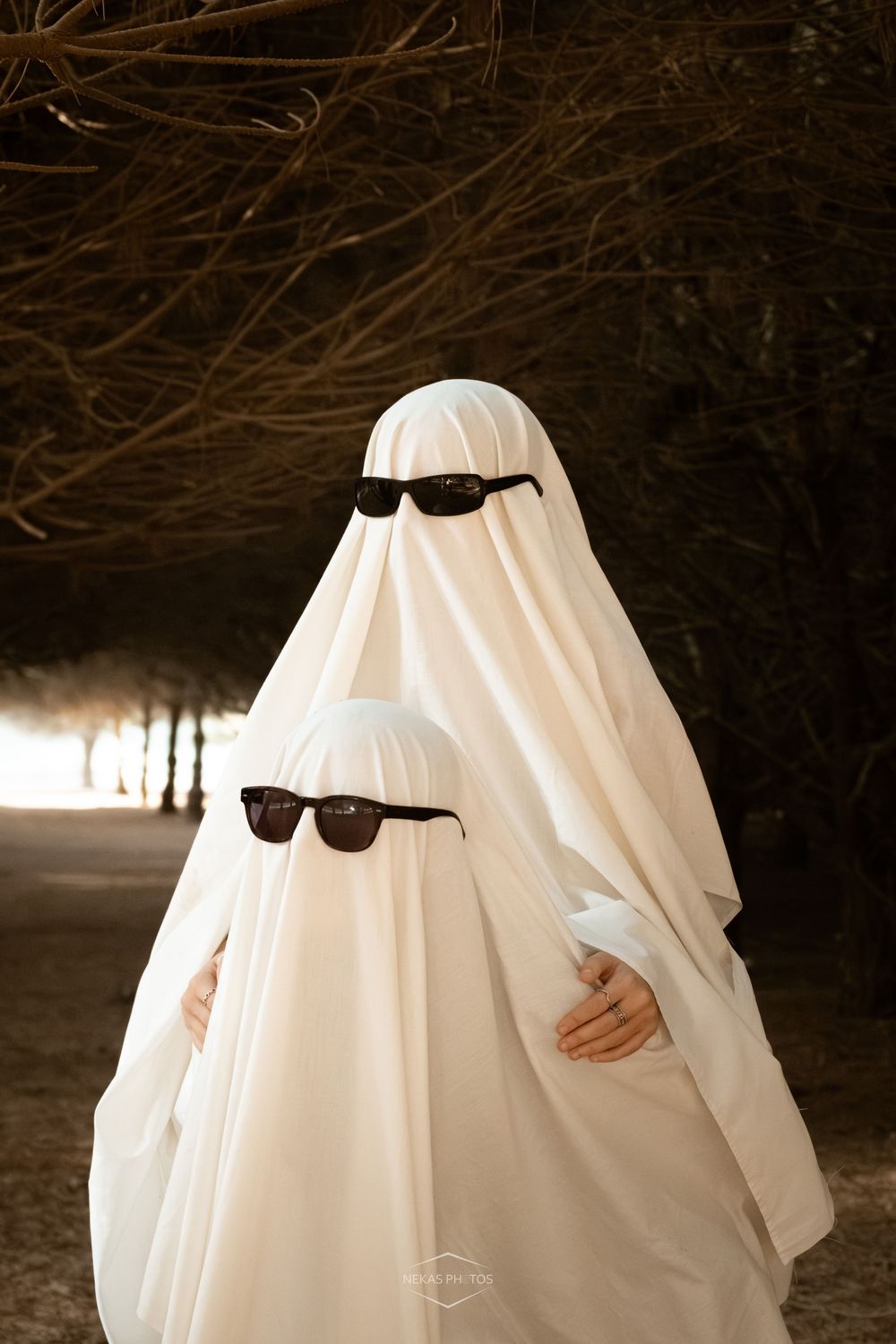 Adult and child in white sheet ghost costumes with black sunglasses posing together in a dark forest path.