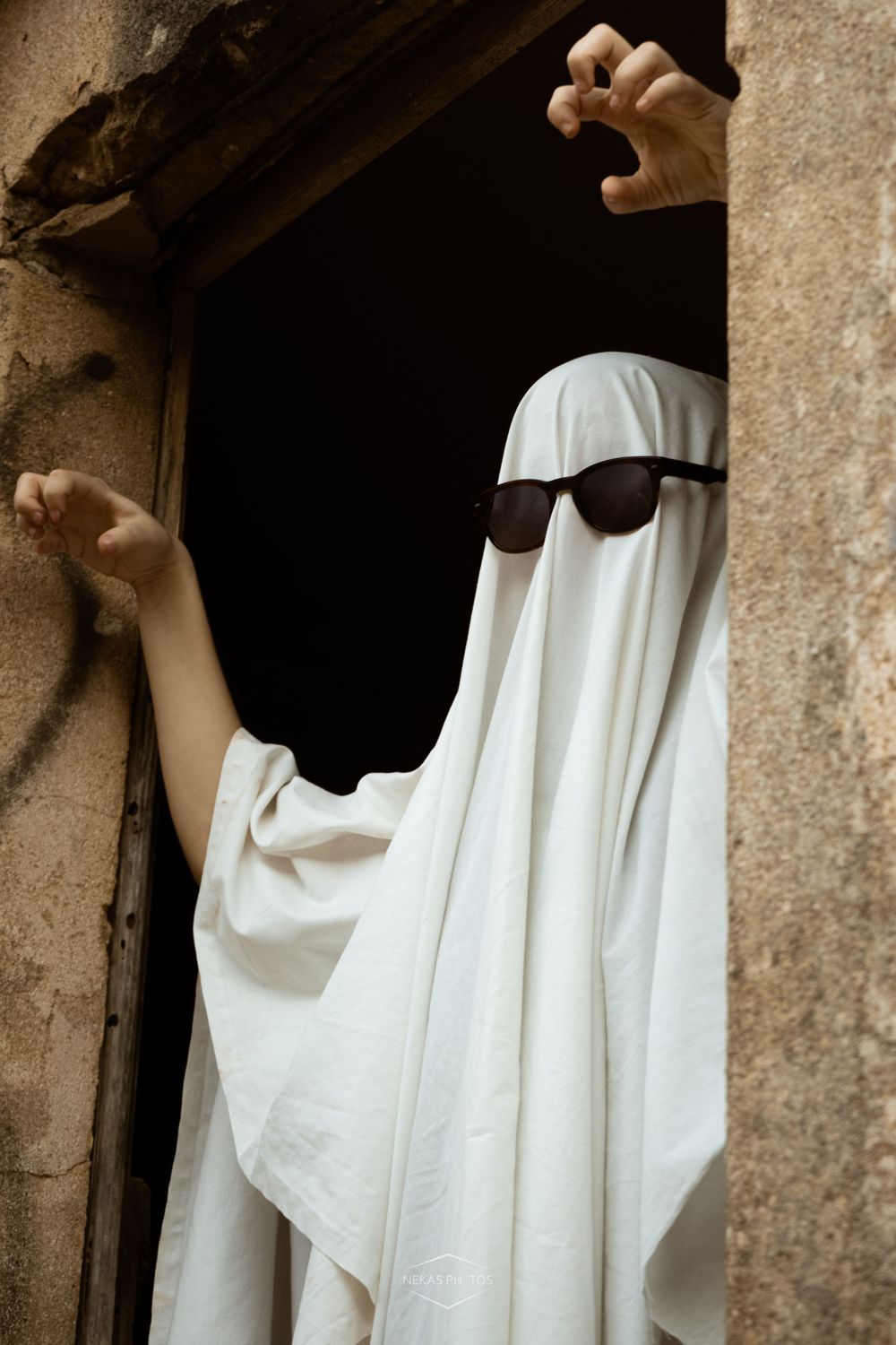 Person in white ghost costume with sunglasses posing in a dark doorway with hands raised in a spooky gesture.