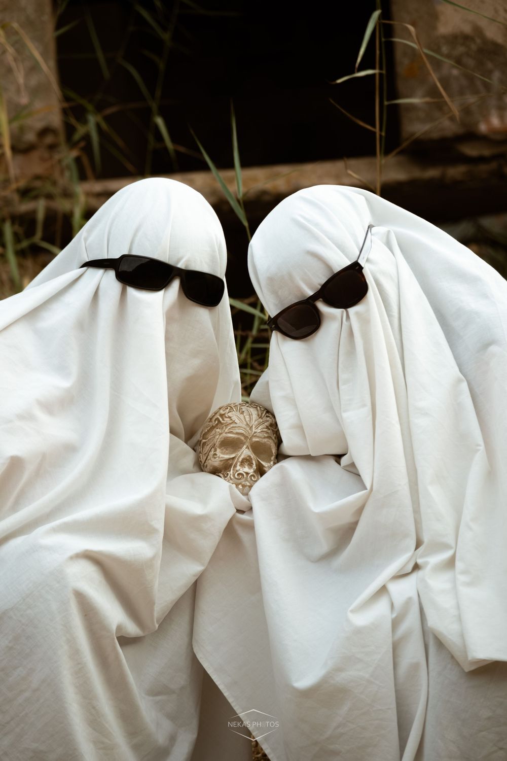 Two people in white ghost costumes with sunglasses holding a skull decoration for Halloween.