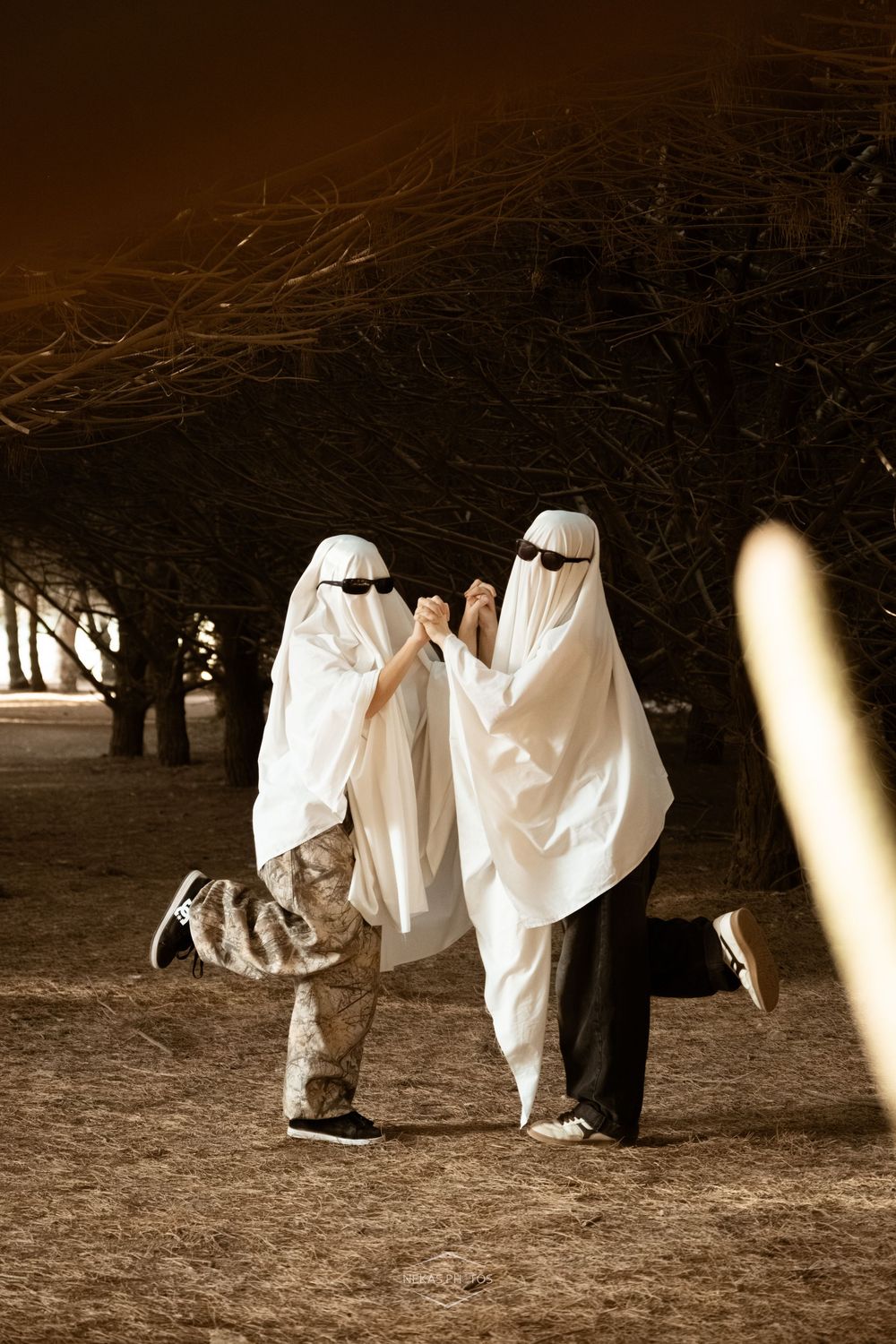 Two people in white ghost Halloween costumes with sunglasses hold hands dancing outdoors at night near bare trees.