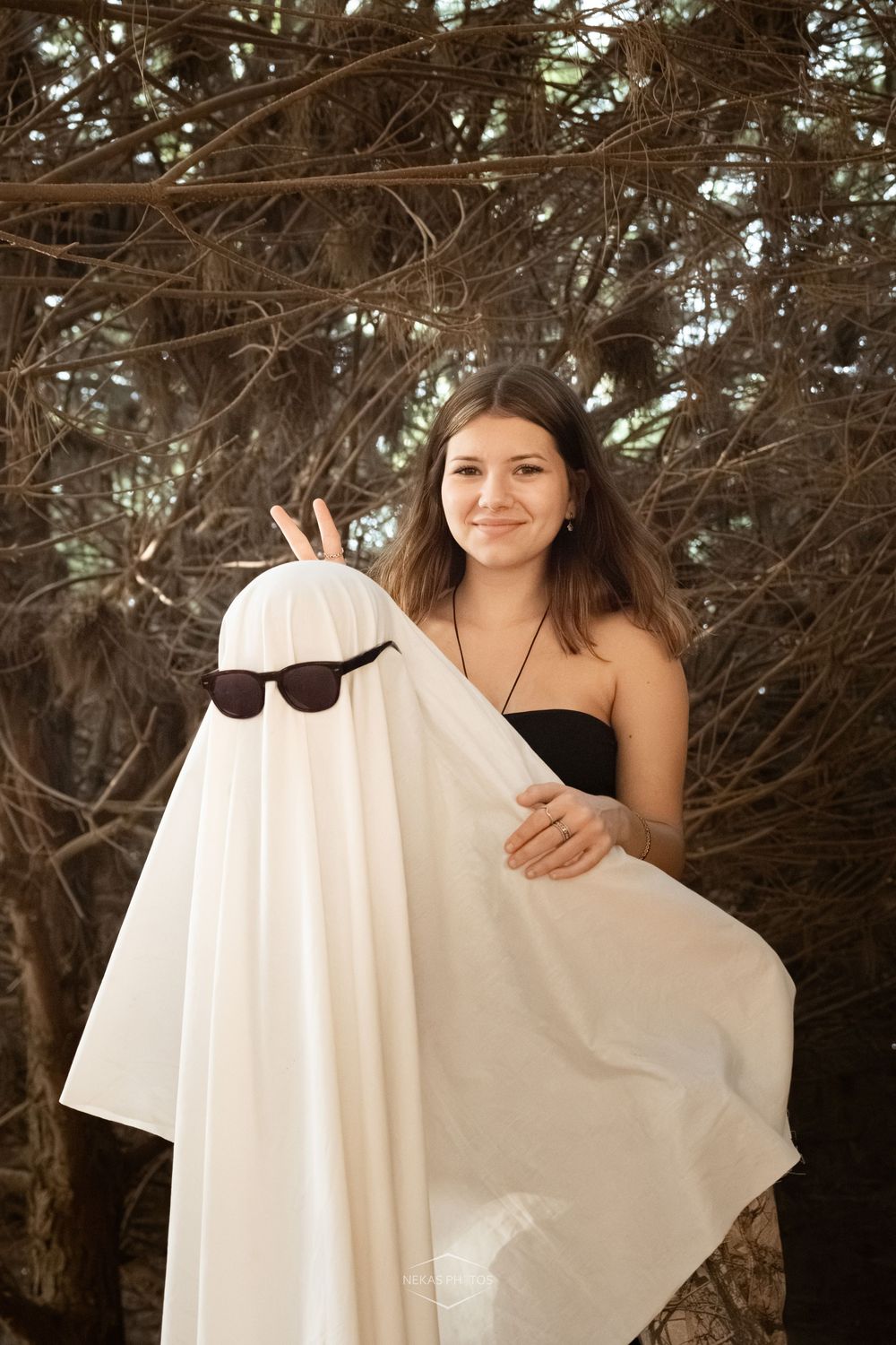 Young woman smiling outdoors holding a white sheet ghost costume with sunglasses in a spooky forest setting.