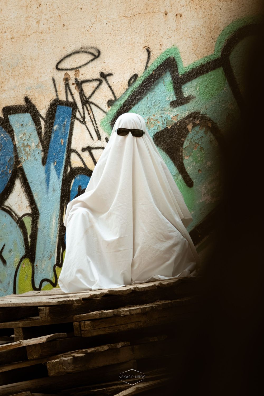 Person in white ghost costume with sunglasses sitting on wooden pallets in front of colorful graffiti wall.