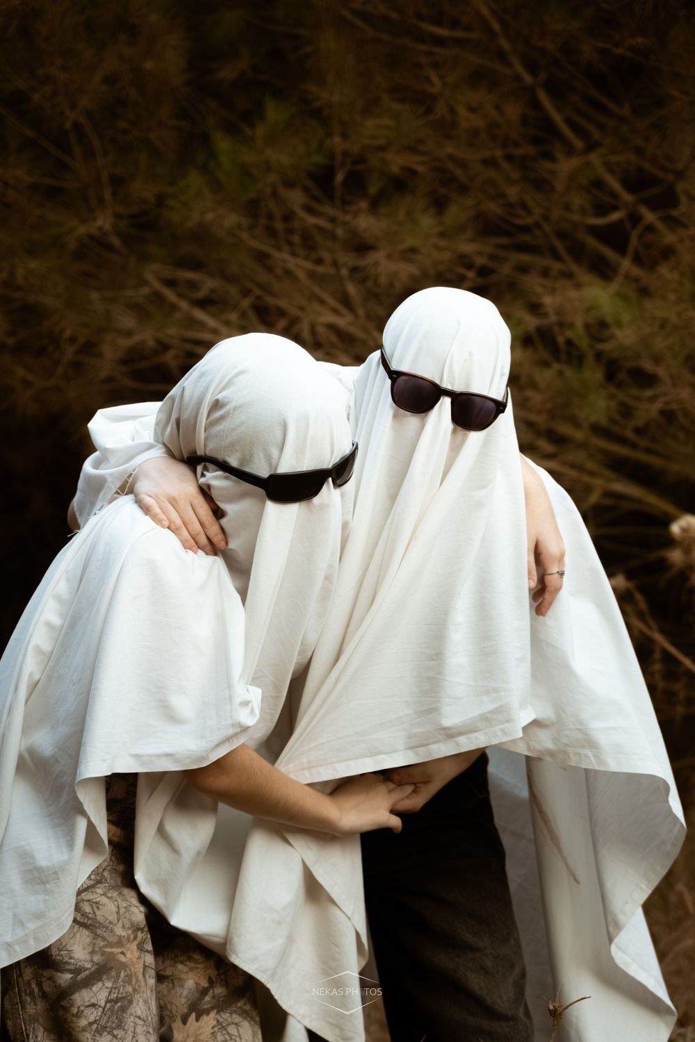 Two people dressed as ghosts in white sheets and sunglasses hugging outdoors near dark green foliage.