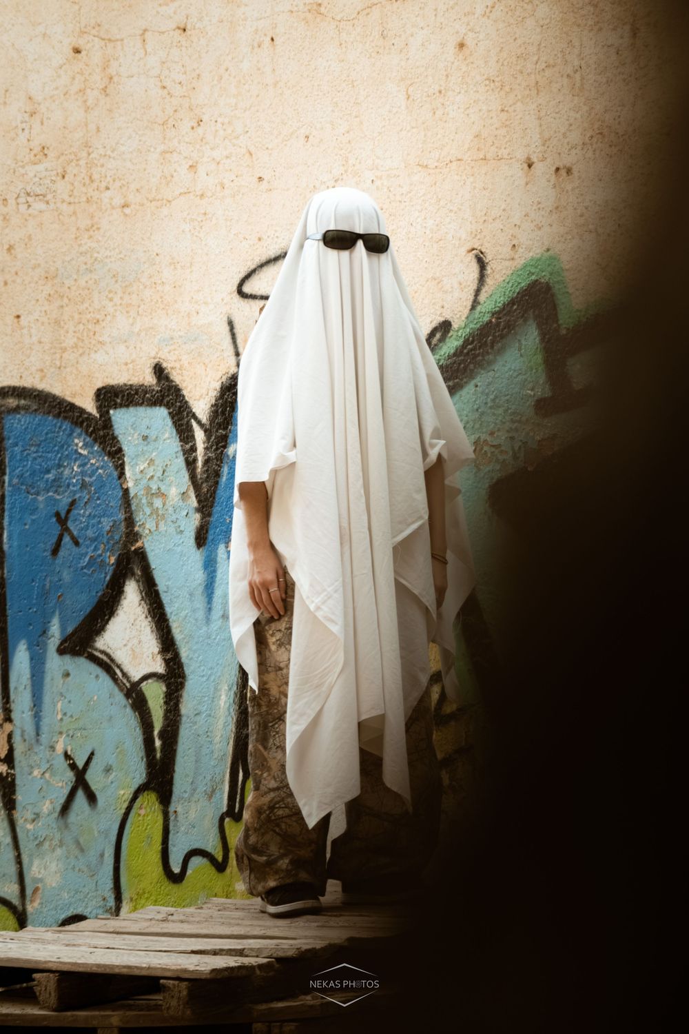 Person dressed as ghost with sunglasses standing near graffiti wall on wooden pallet.