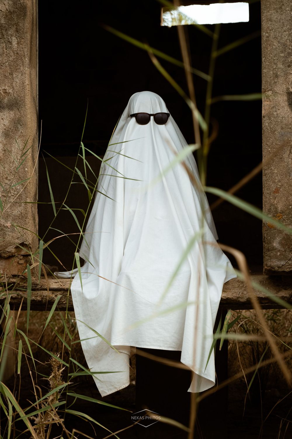 Person in white ghost costume with sunglasses standing in abandoned building doorway surrounded by tall grass.