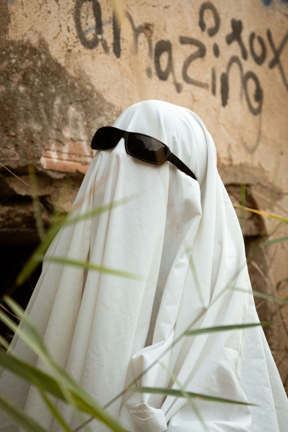 Person in white ghost costume with black sunglasses posing against a graffiti wall outdoors.