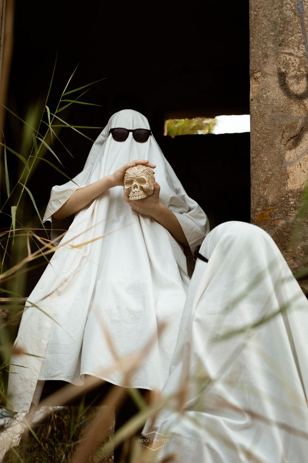 Person in white ghost costume with sunglasses holding a skull prop outdoors for Halloween.
