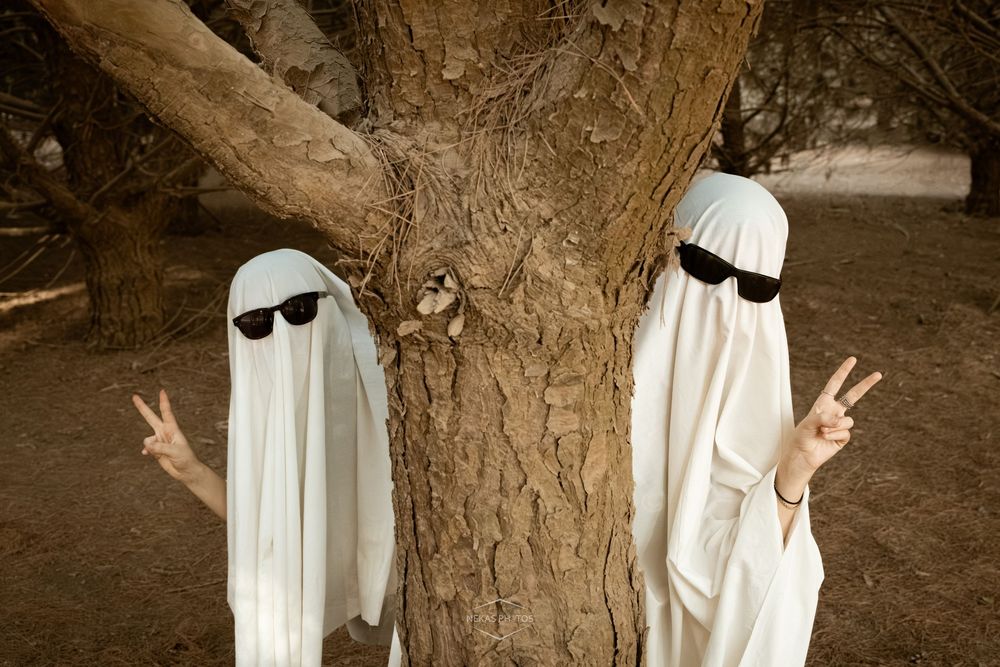 Two people in white ghost costumes with sunglasses making peace signs beside a tree.