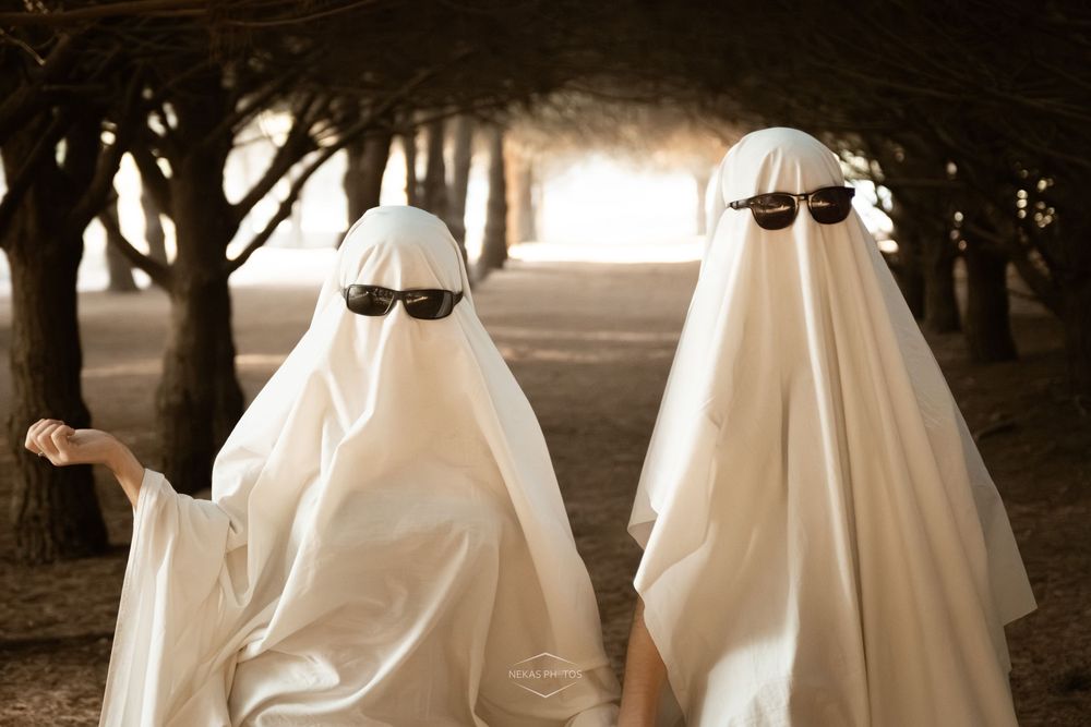 Two people in white ghost costumes wearing sunglasses walk through a tree-lined path on Halloween.