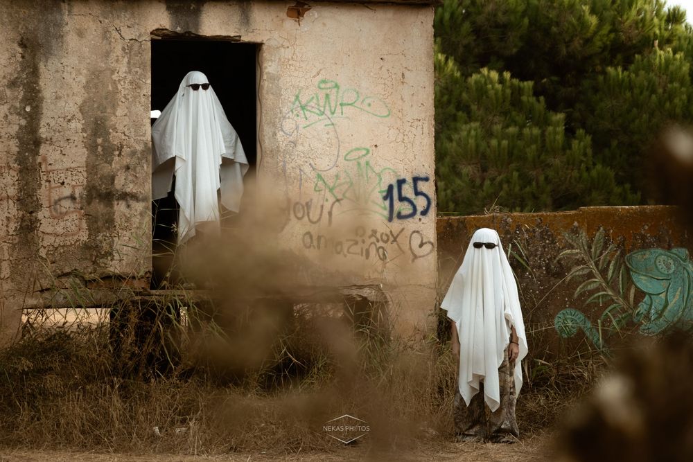 Two people dressed as ghosts in white sheets near an abandoned graffiti-covered building outdoors.
