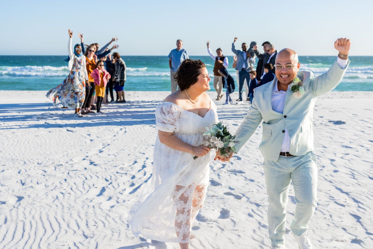 Joyful newlyweds celebrate on a white sand beach with cheering guests behind them, bride holding bouquet, groom in light suit.