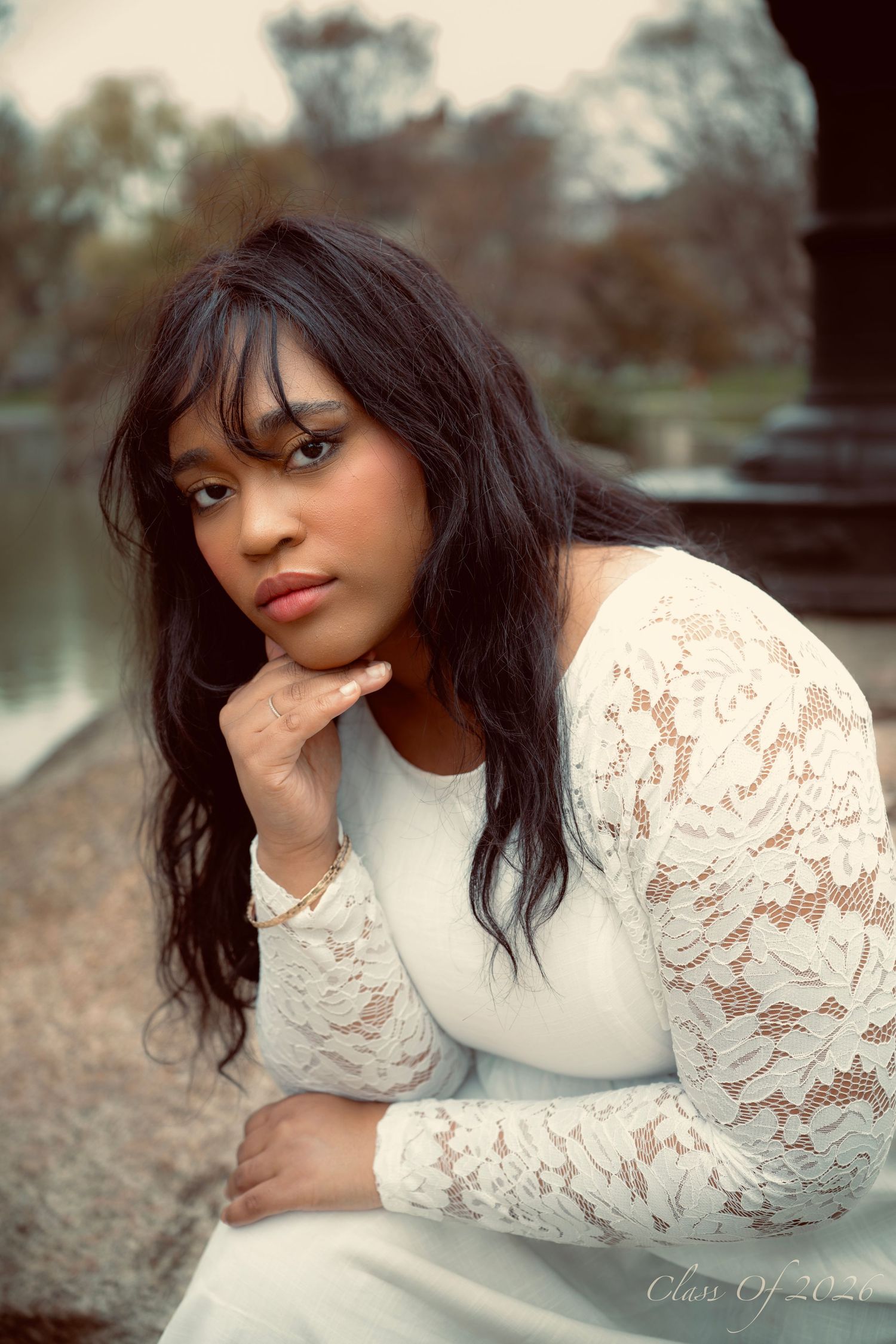 Young woman in white lace dress posing thoughtfully by waterside in soft vintage-style portrait.