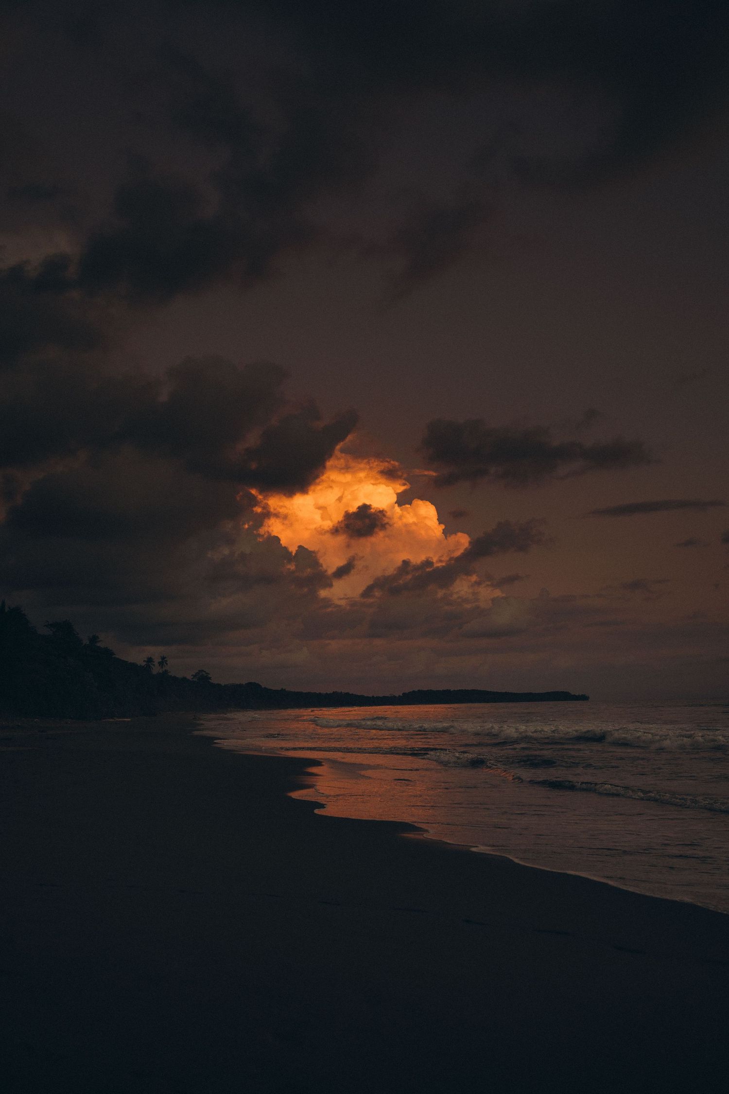 Dramatic beach sunset with glowing orange clouds reflected on wet sand along a dark tropical shoreline.