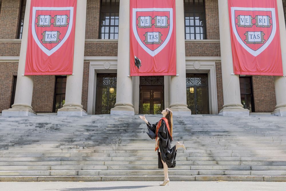 Graduate tosses cap in front of Harvard University's iconic red banners and grand stone steps.