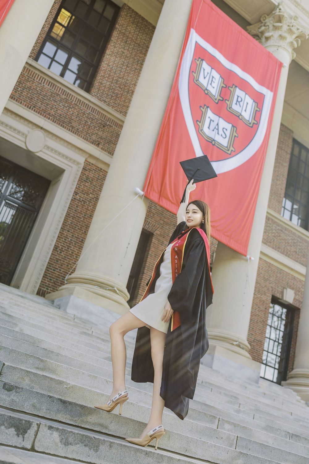 Harvard graduate in cap and gown poses on steps holding mortarboard beneath crimson VERITAS banner.