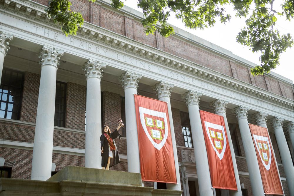 Harvard University building with red banners and white columns, two people standing on steps taking a selfie.