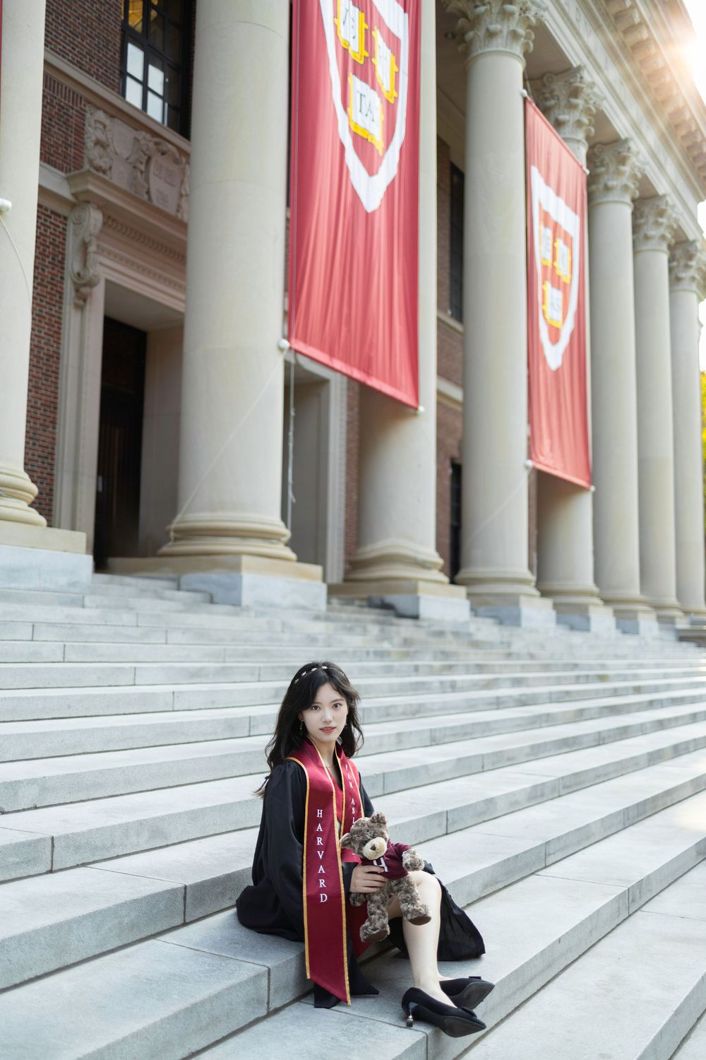 Harvard graduate in black gown and crimson sash sits on steps of Widener Library with school banners.