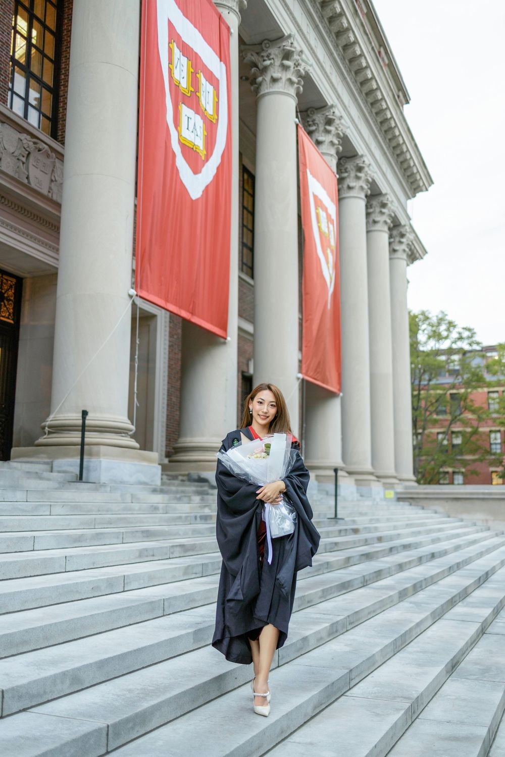 A graduate in cap and gown holds flowers on the steps of Harvard University, with red banners in the background.