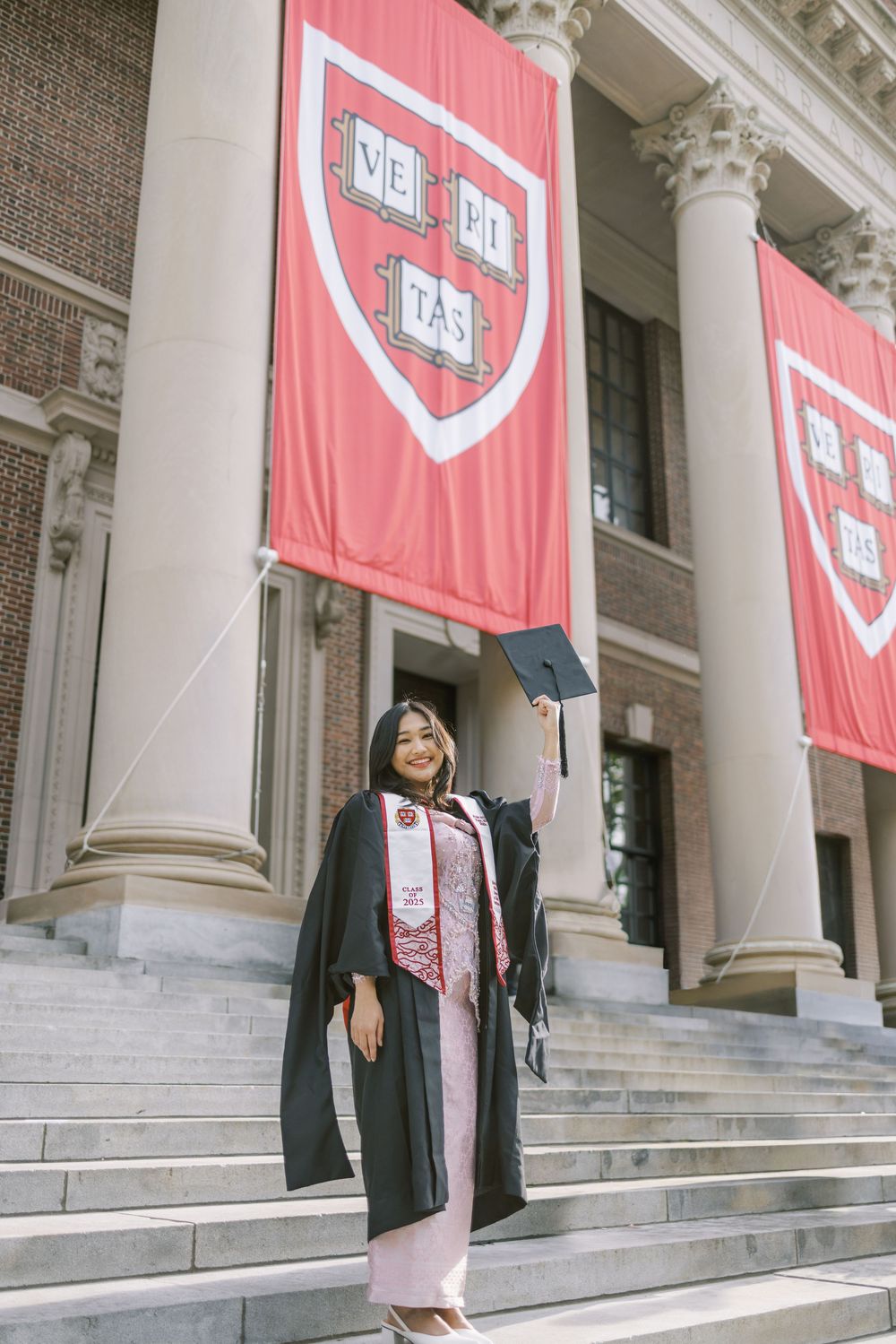 Harvard graduate in cap and gown poses on university steps with crimson Veritas banners behind her.
