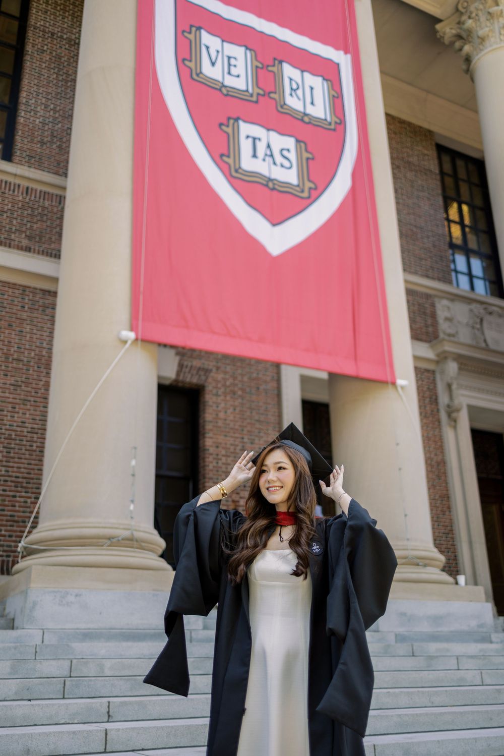 Harvard graduate in cap and gown poses proudly on steps beneath the iconic Veritas shield banner.