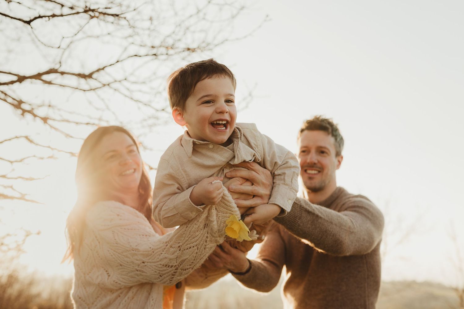 Happy family with parents lifting laughing toddler boy outdoors in warm golden sunset light during autumn.