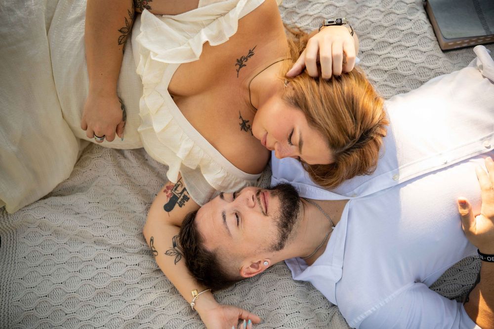A tattooed couple sharing a romantic moment lying on a bed, with a woman leaning over a smiling man.