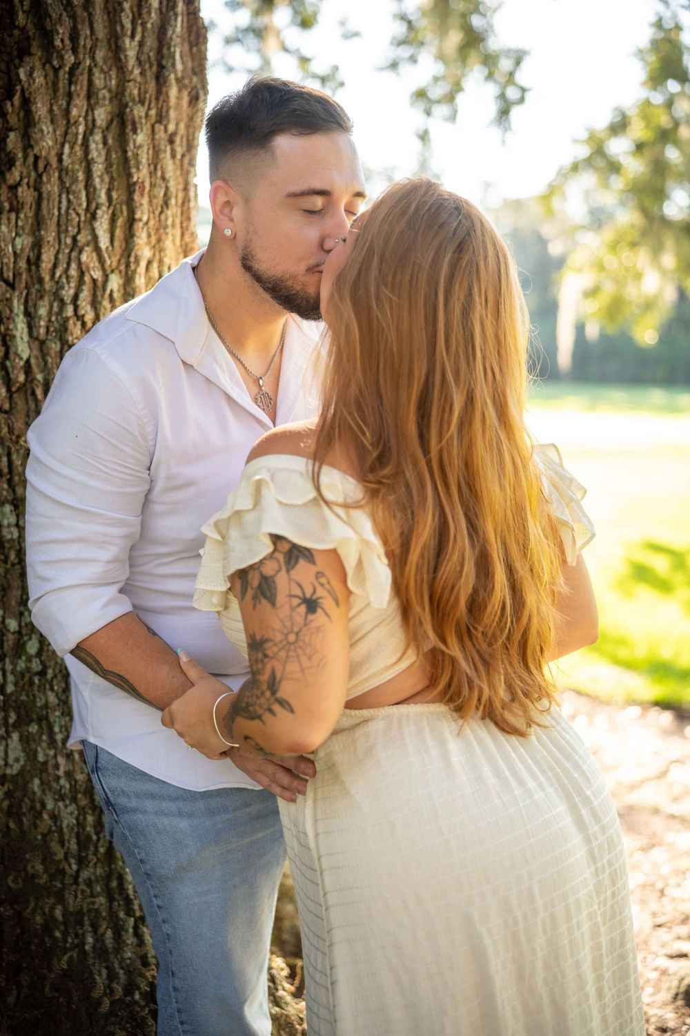 Couple sharing a romantic kiss against a tree outdoors, woman in cream ruffled dress with tattoos, man in white shirt.