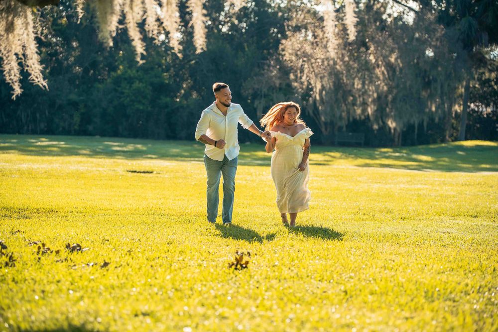 A joyful couple runs hand-in-hand through a sunlit green meadow surrounded by lush trees on a bright sunny day.