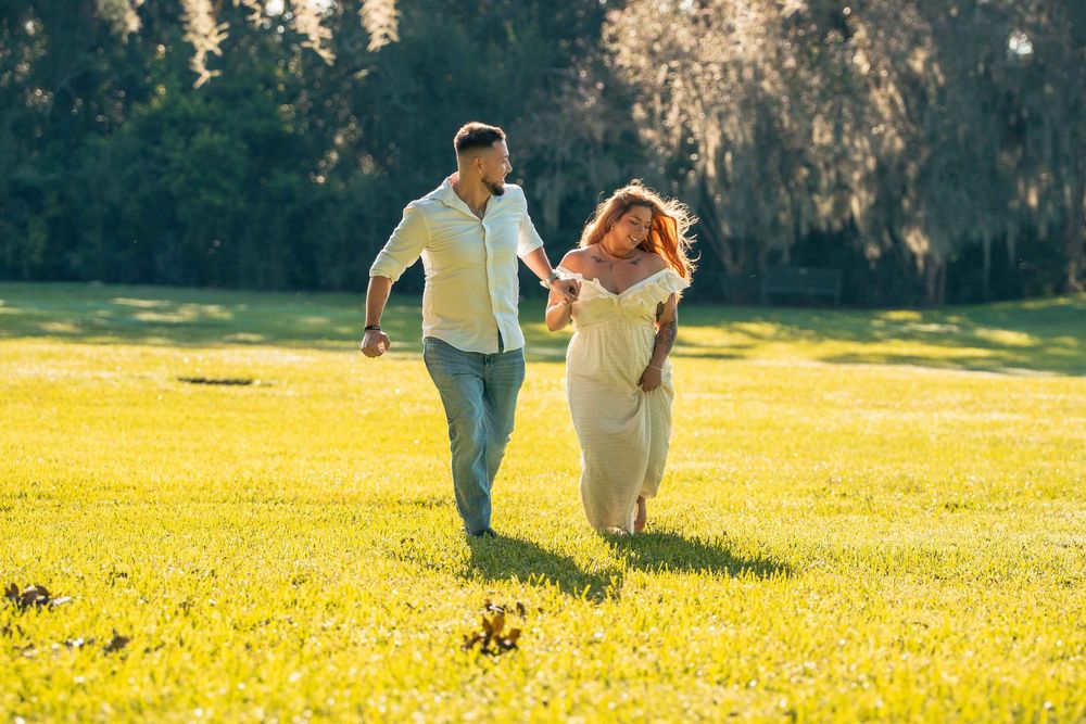 A happy couple holds hands while running through a sunny green park, with the woman wearing a flowing white dress.