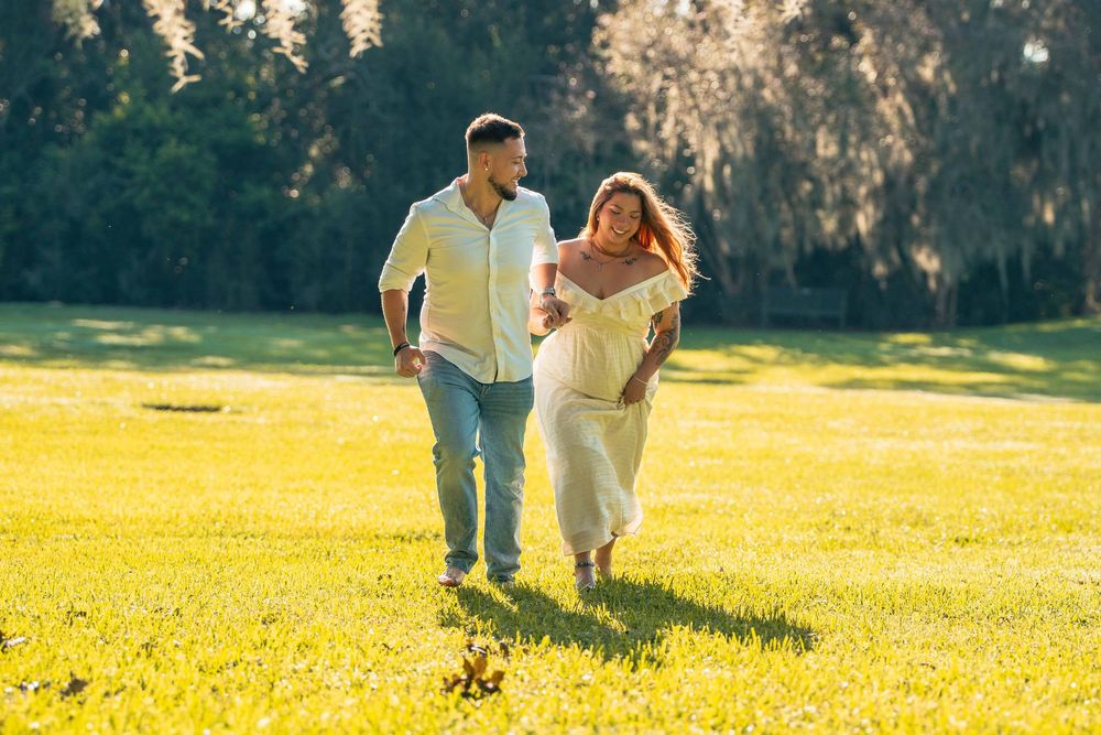 Pregnant couple walking hand in hand on sunny green lawn, woman in white dress, man in light blue jeans and shirt.