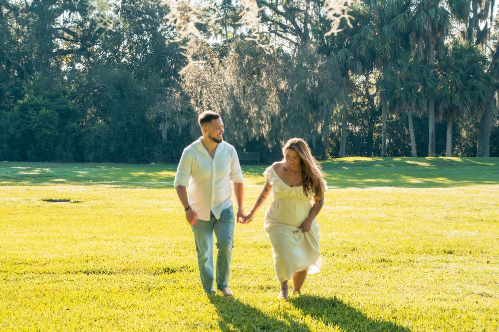 Couple holding hands walking through a sunny green park with Spanish moss-draped trees, woman in yellow maternity dress.