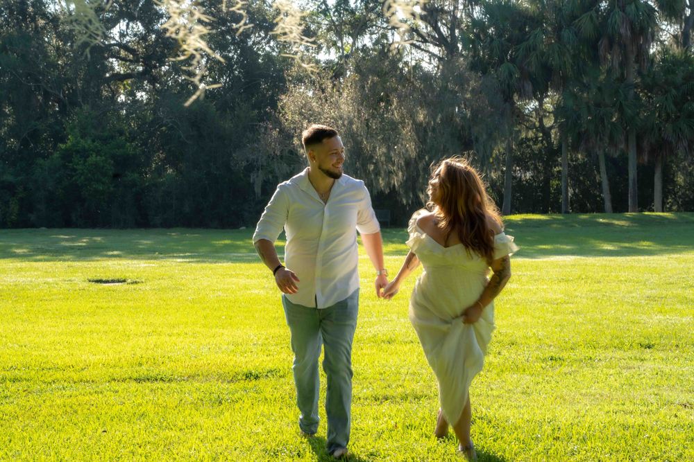 Couple holding hands and walking together on lush green grass in a sunny park surrounded by tall trees.