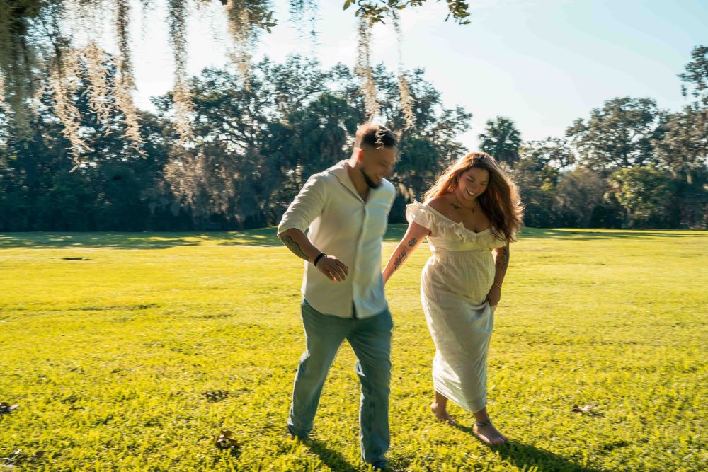 Couple walking hand in hand on lush green grass in a sunlit park, woman in white dress, man in casual attire.