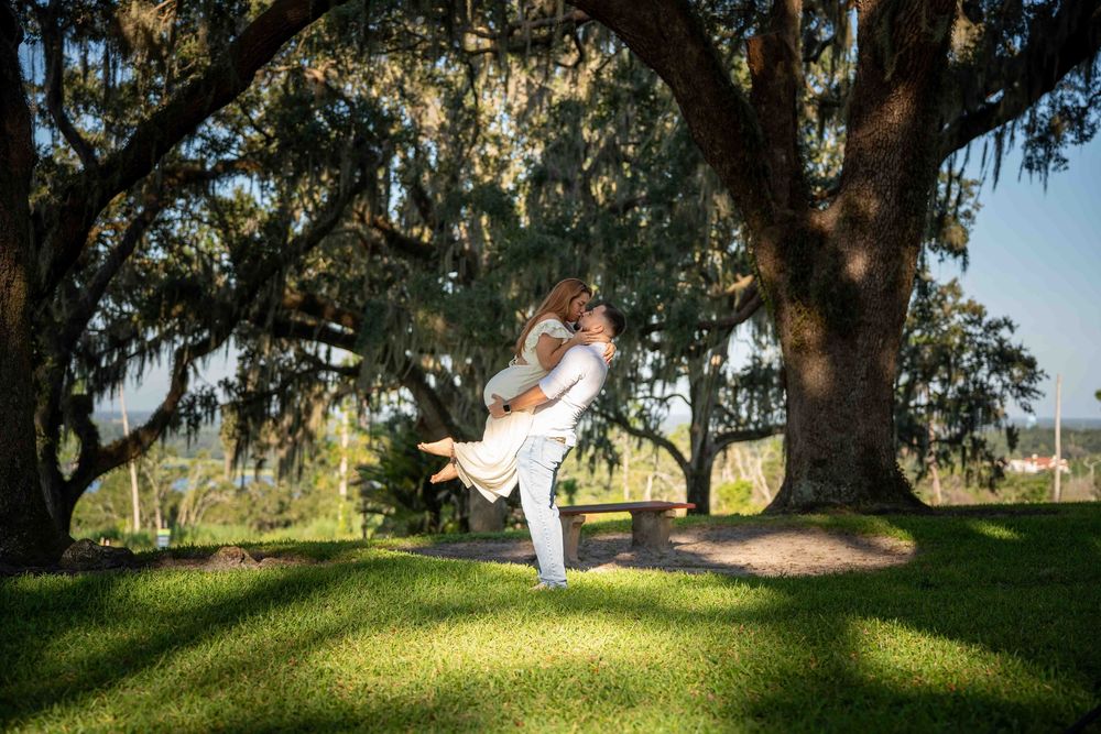 Couple sharing a romantic lift embrace under a sprawling moss-draped oak tree on a sunny green lawn.