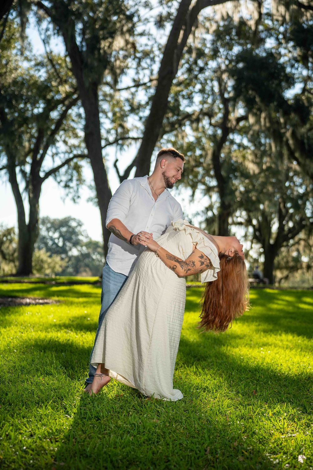 Couple dancing outdoors under moss-draped oak trees on lush green grass, man dipping woman in white dress.